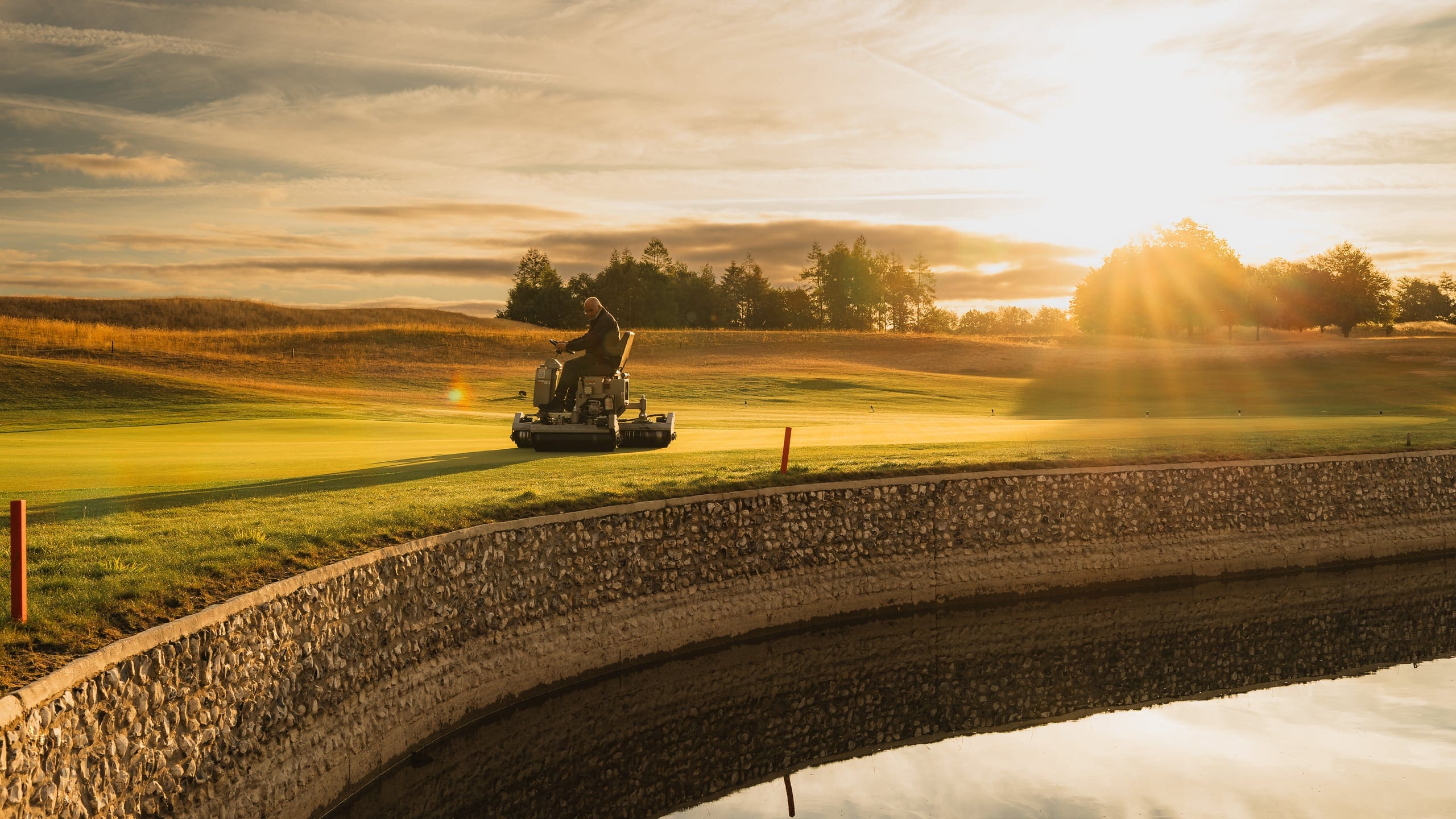 A greenkeeper mowing into a low sun, light flaring across the frame