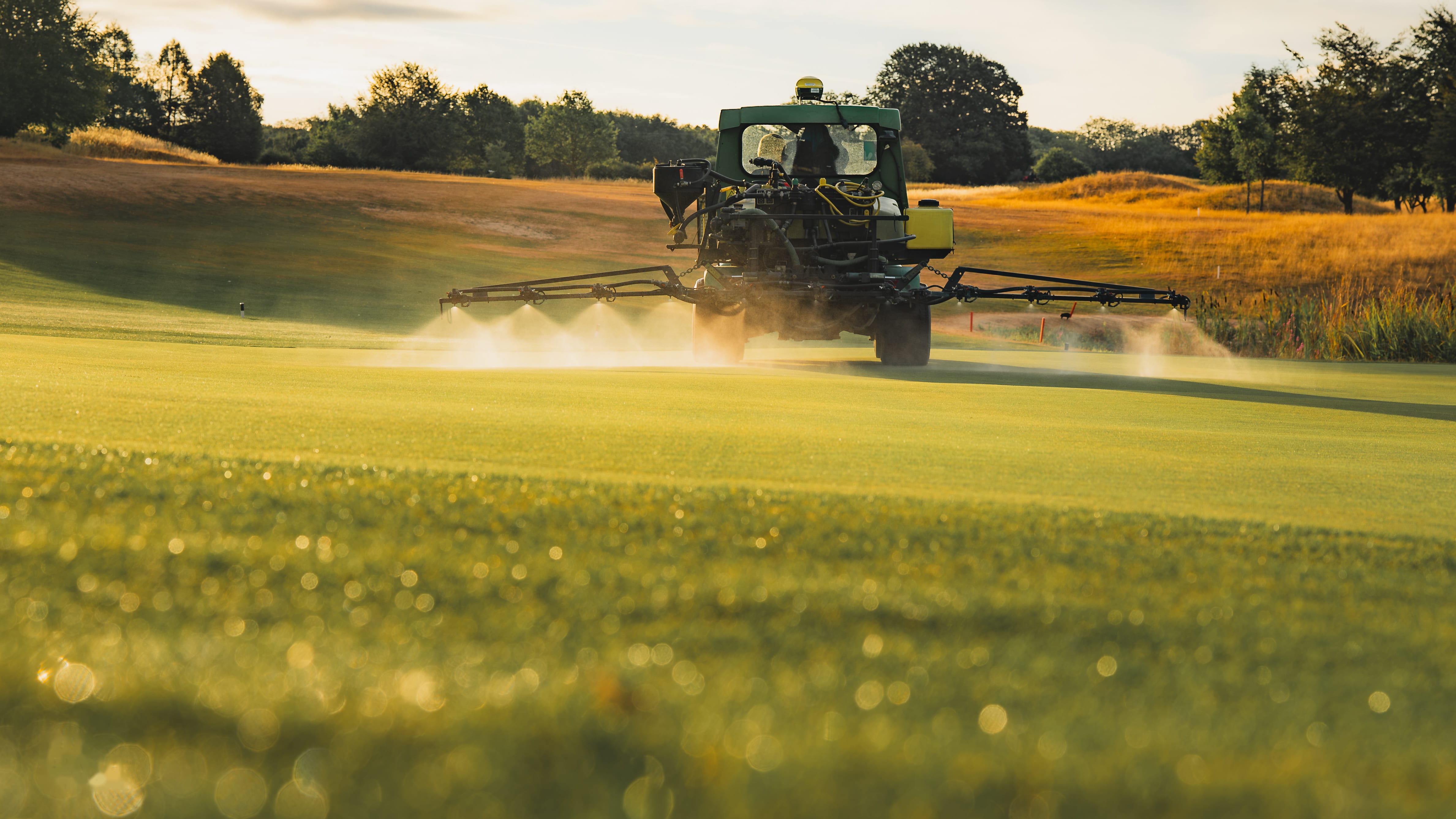 A mower throwing up a fine spray of dew as it cuts, backlit by low sun