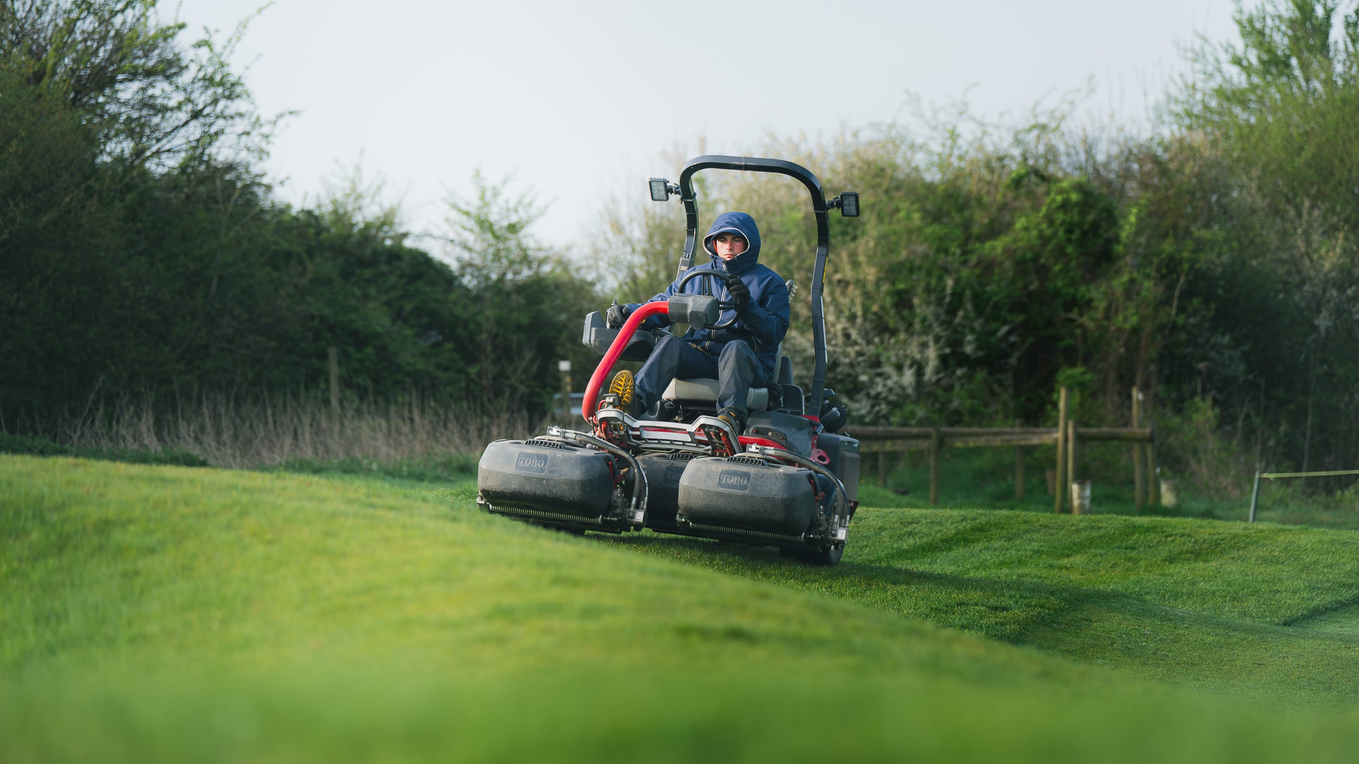 A greenkeeper on a mower approaching along a tree-lined fairway