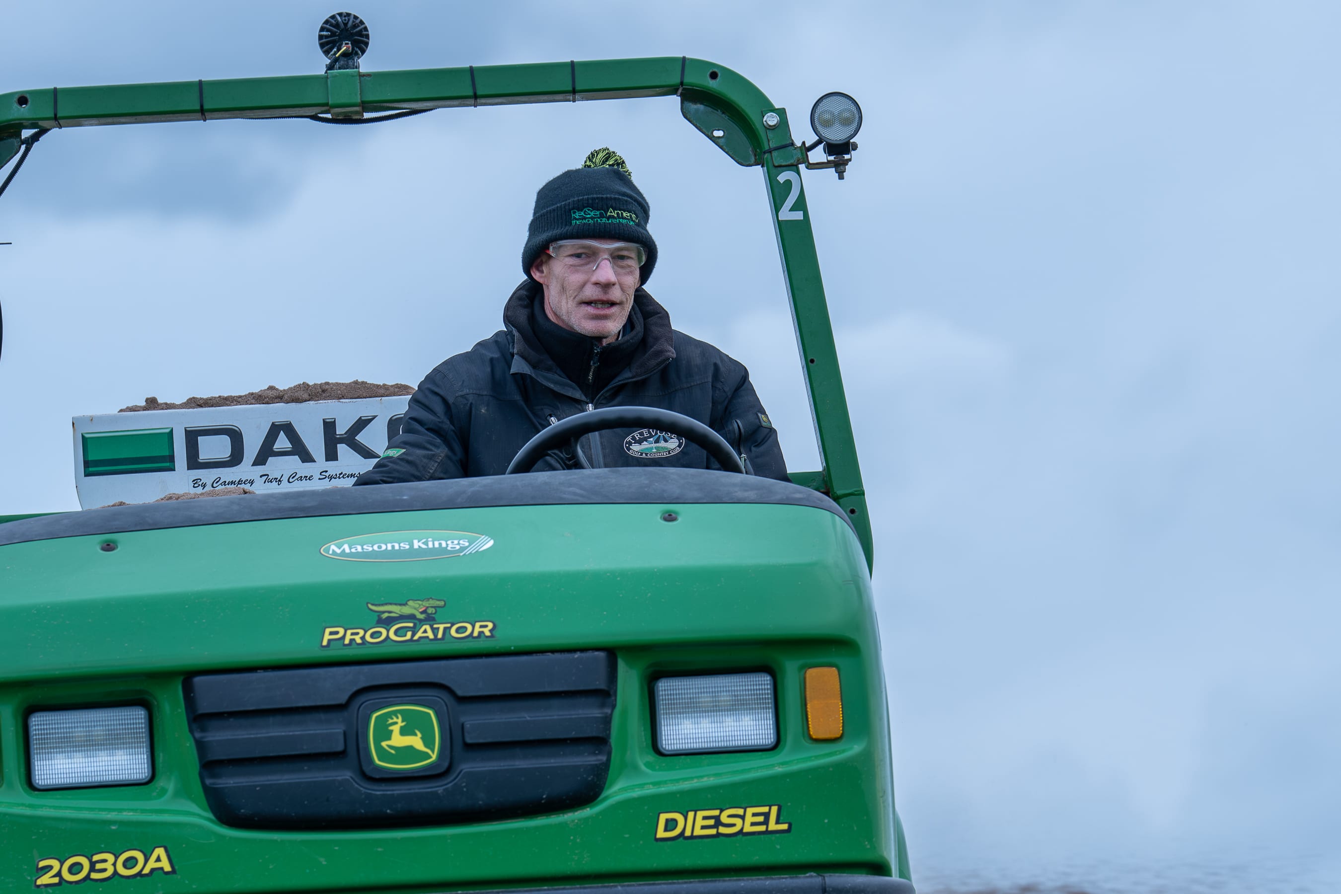A greenkeeper at the wheel of a John Deere ProGator carrying a top dresser, the Masons Kings decal on the bonnet