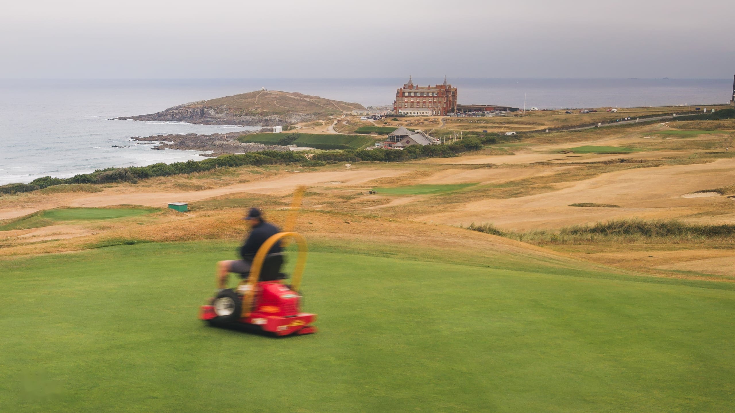 A greenkeeper mowing a green above the sea, the headland hotel beyond