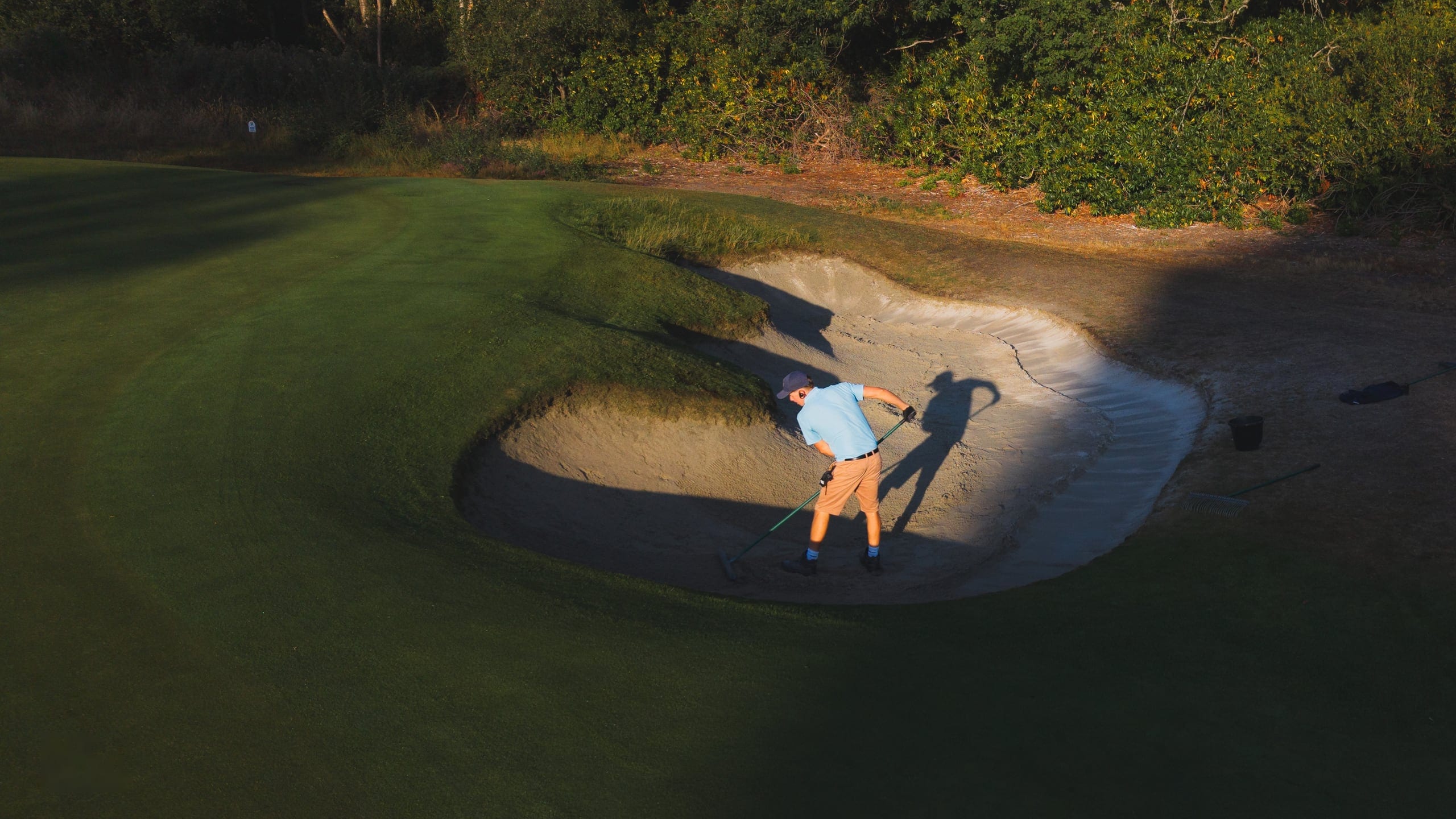 A greenkeeper raking a bunker at low sun, seen from directly above, his shadow stretched across the sand