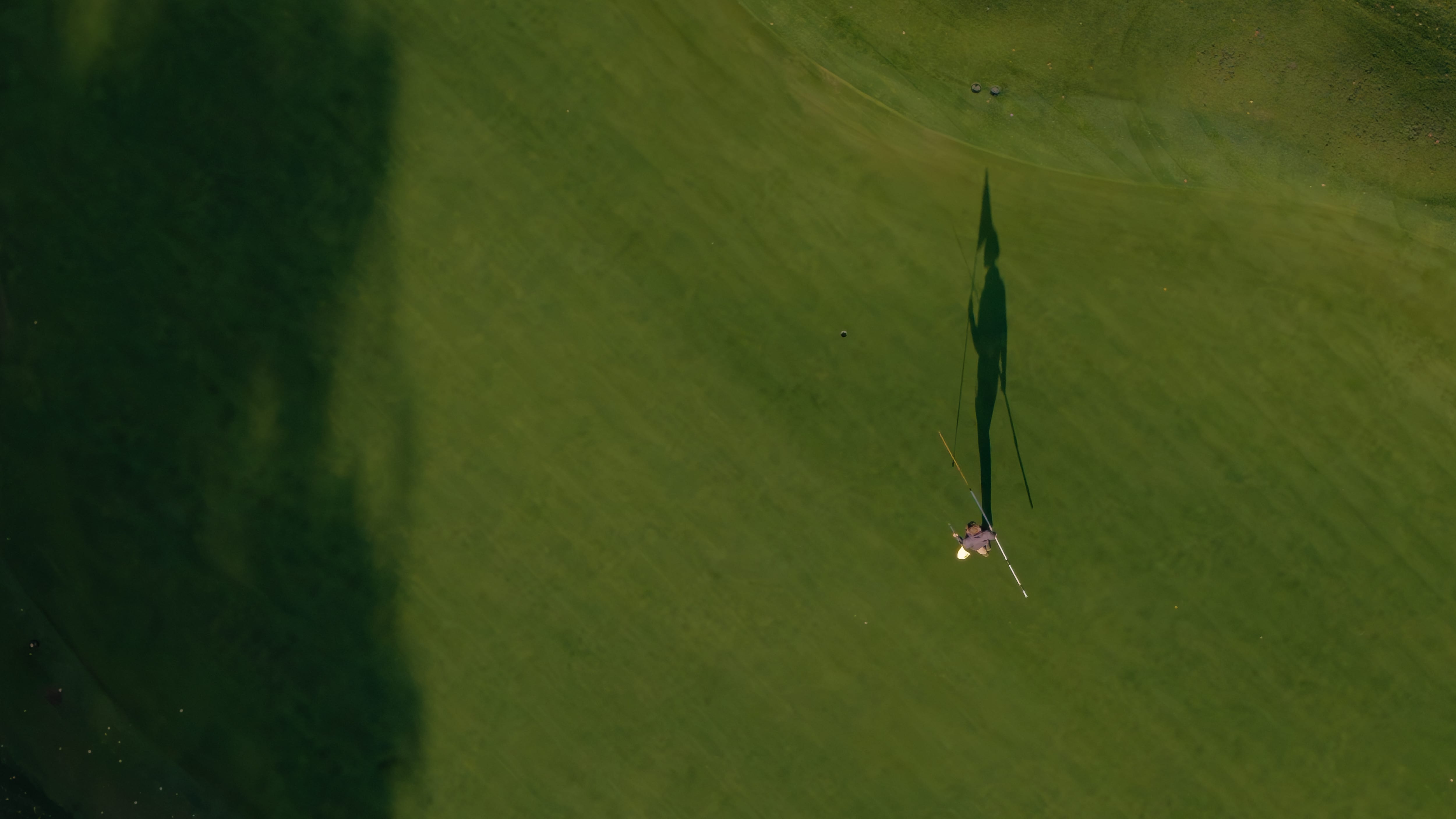 A golfer putting on a green at low sun, seen from directly above, his shadow and the flagstick stretched across the surface
