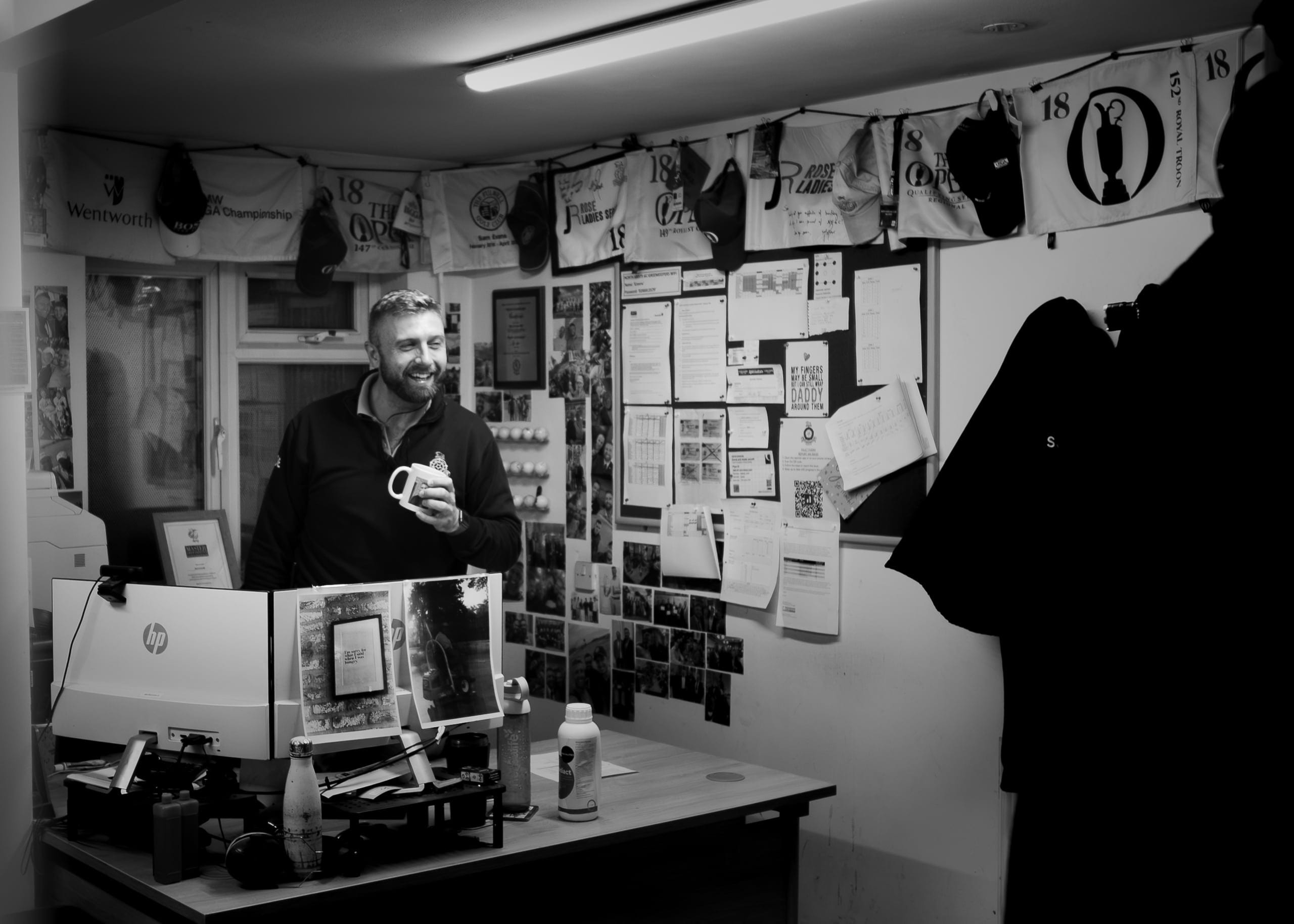 A greenkeeper laughing over a mug of tea in the maintenance office, tournament flags and photographs pinned to the wall behind him