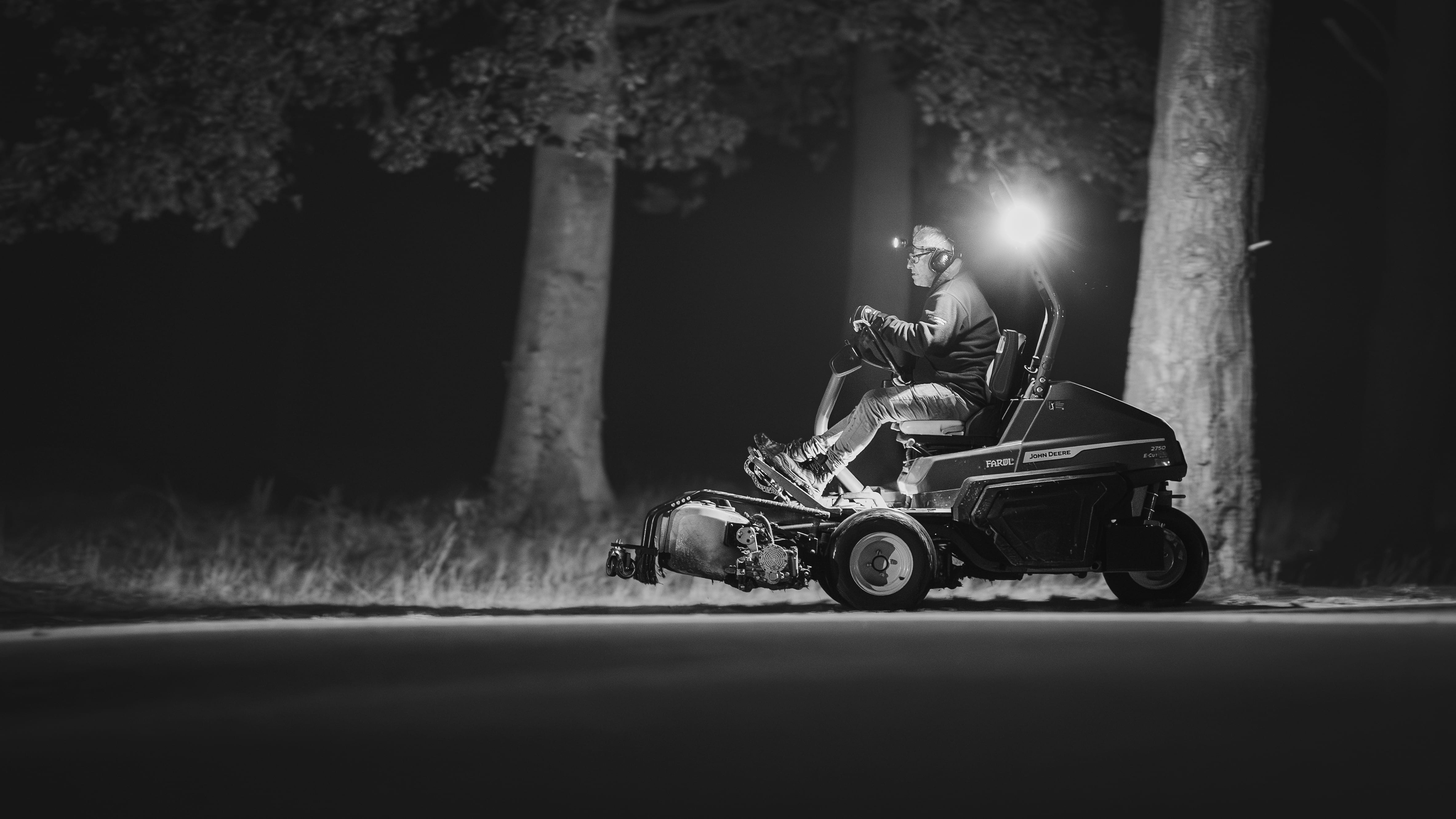 A greenkeeper mowing between trees after dark, lit by the machine's own lamps, in black and white