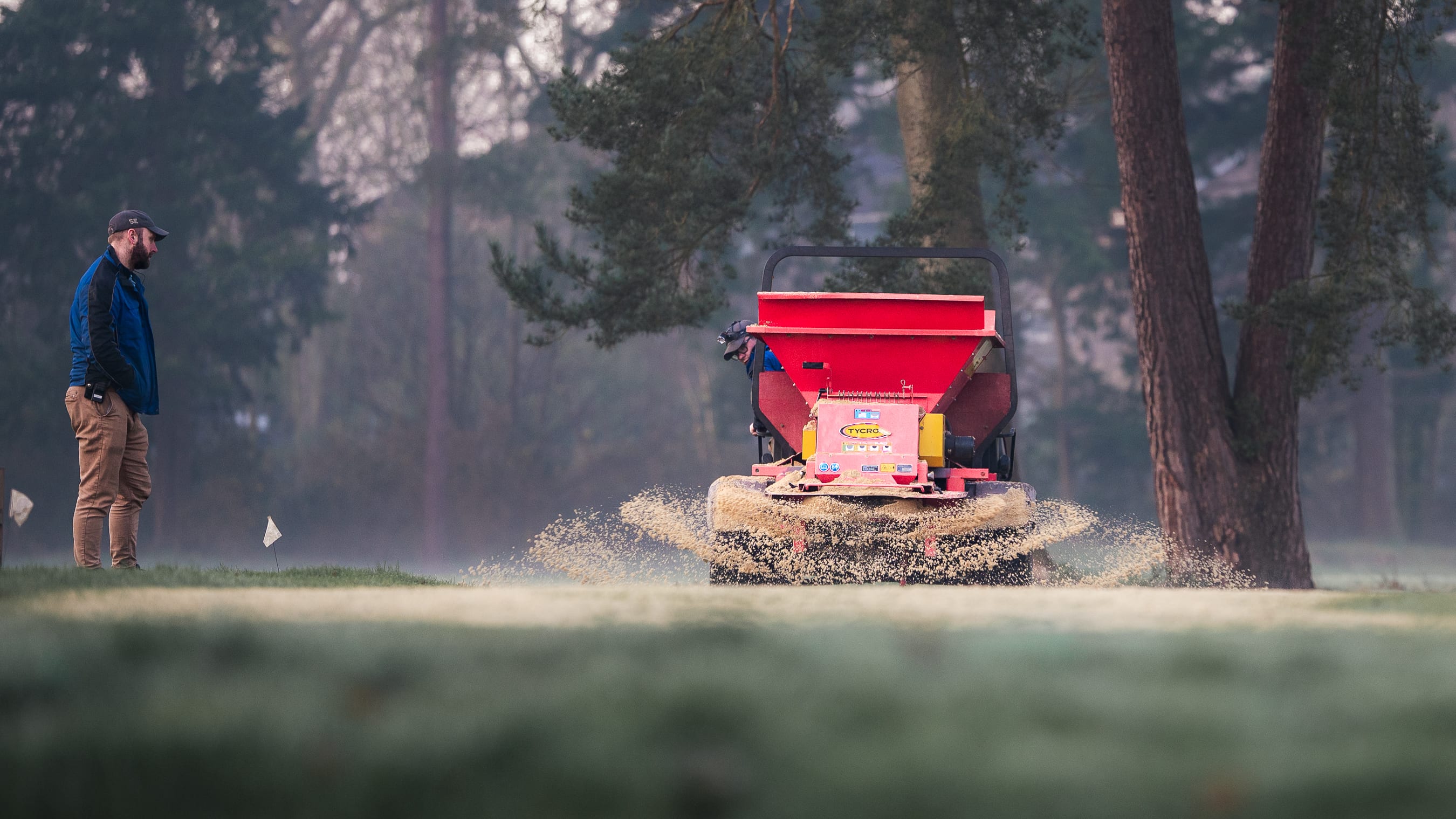 A spreader throwing fertiliser across a tee in the mist, the operator watching from the edge