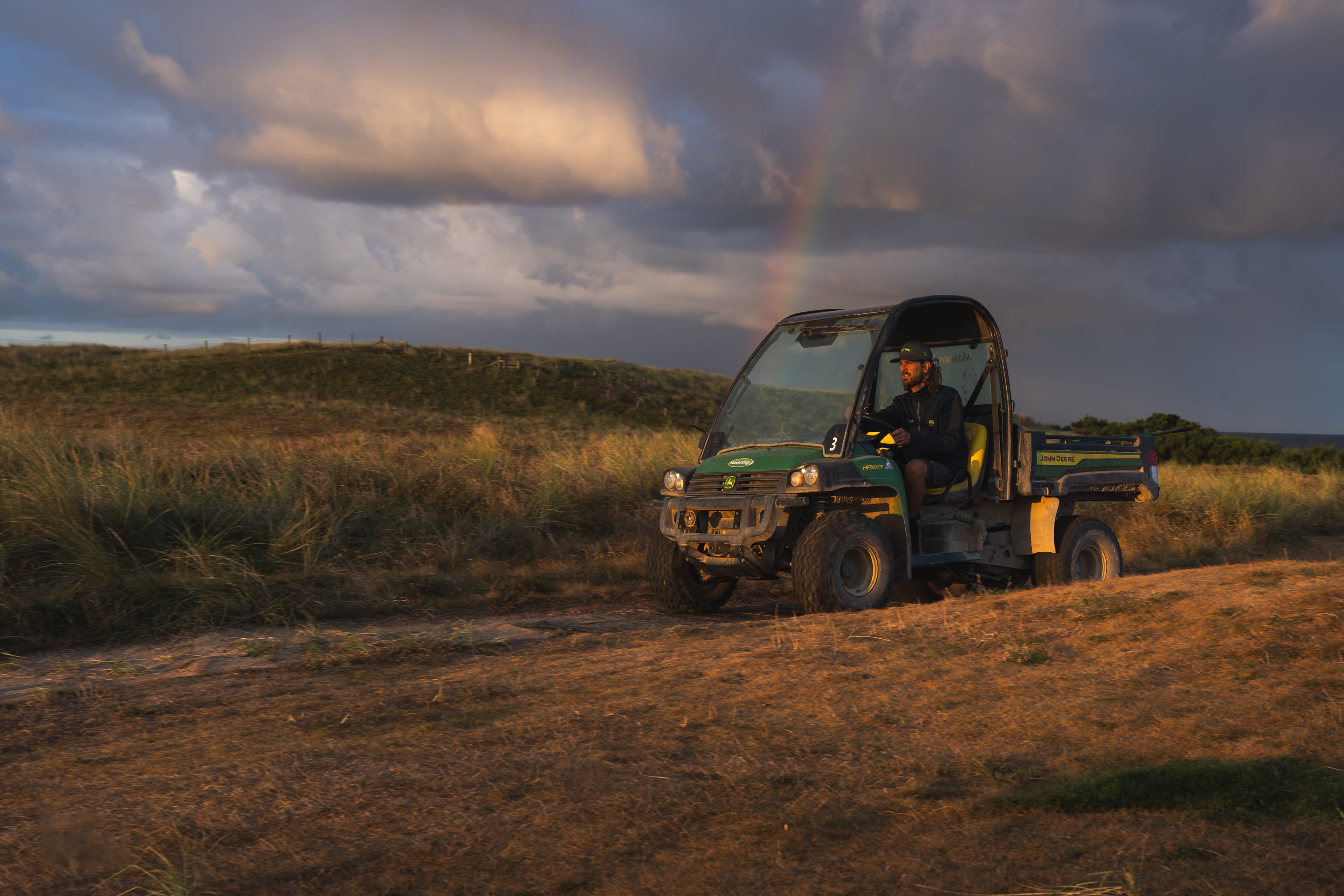 A greenkeeper driving a utility buggy under a rainbow at sunset