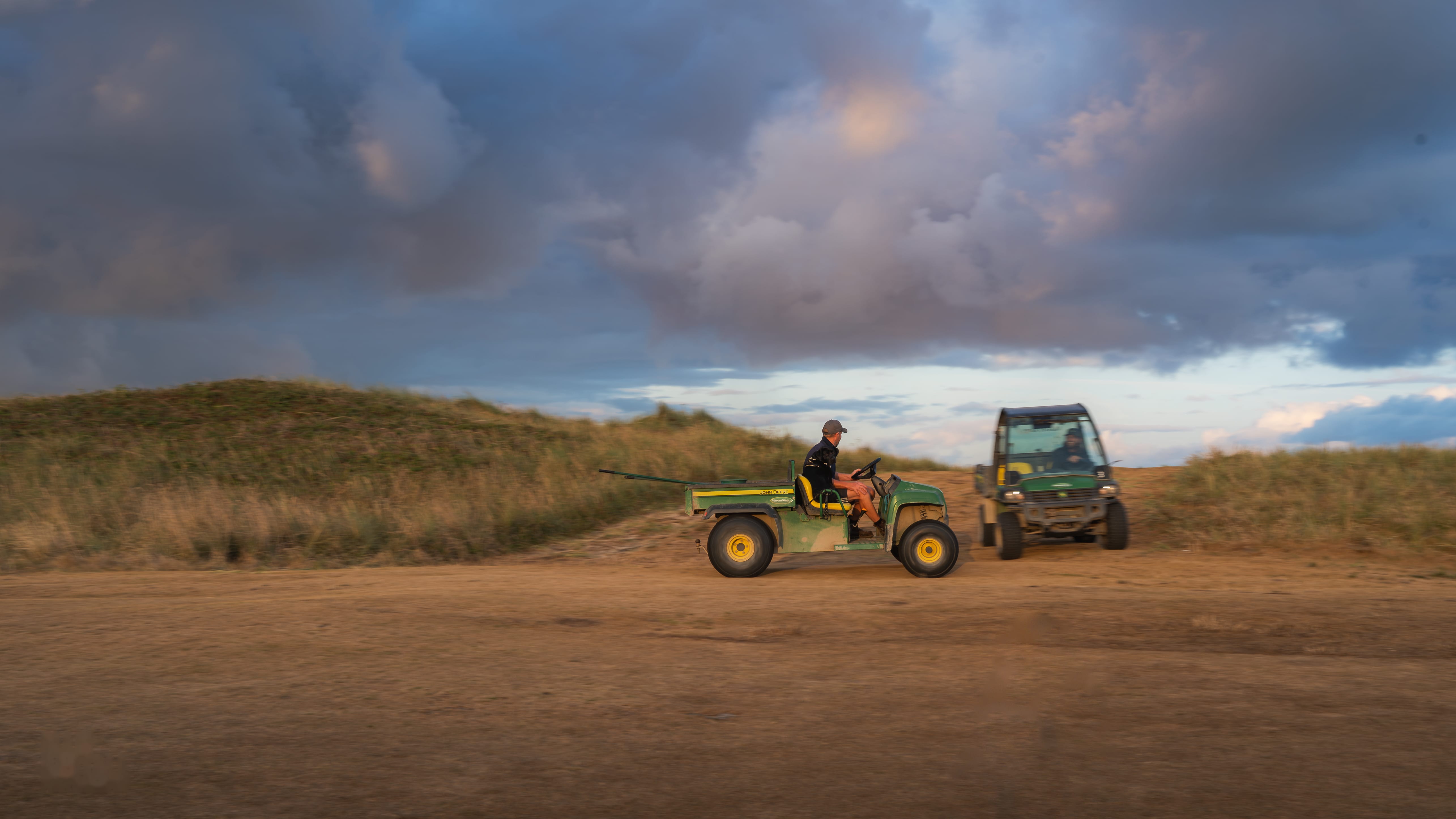 A greenkeeper towing a mower along a sand track through the dunes under a heavy sky