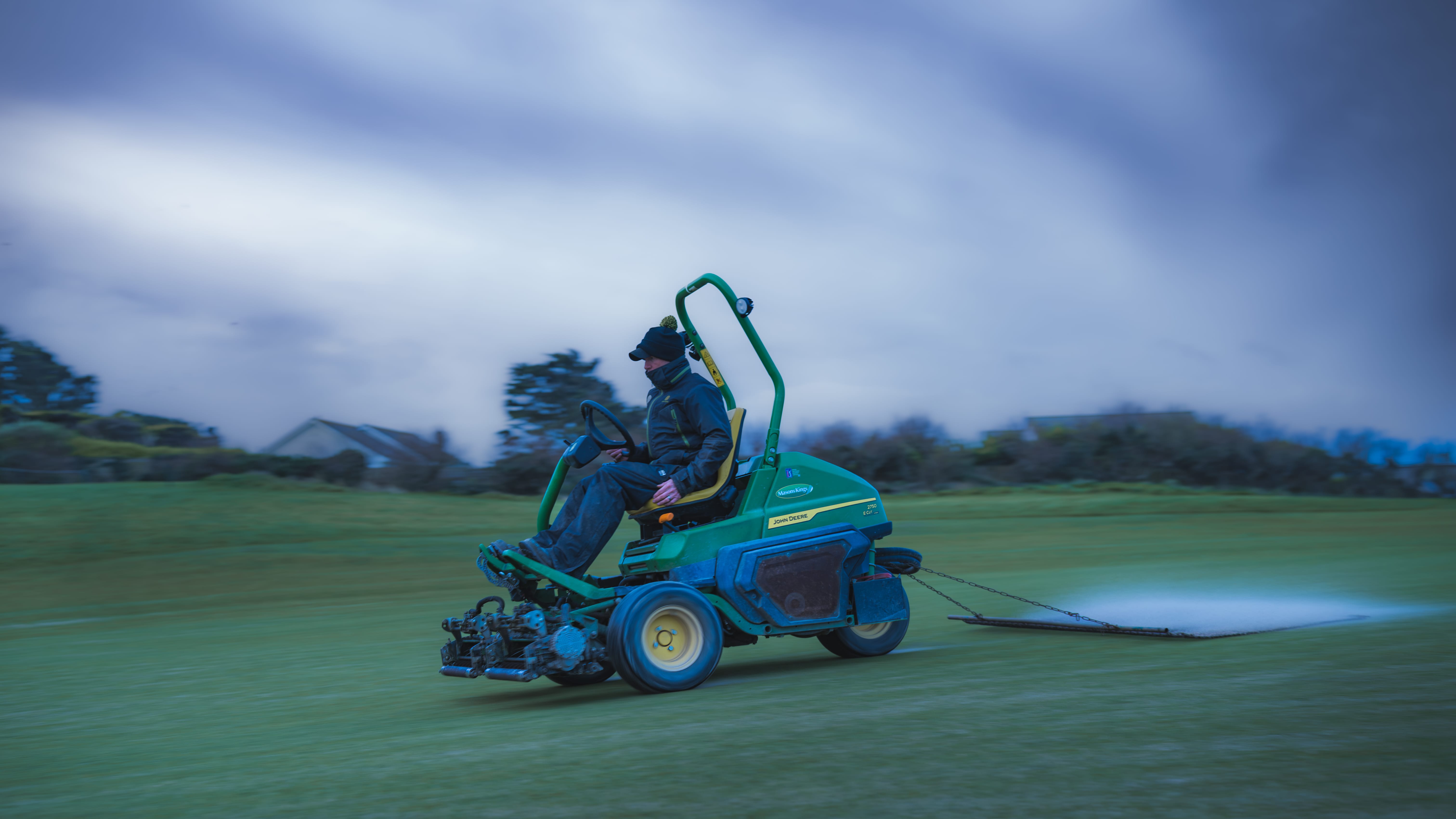 A greenkeeper mowing a green at dusk, headlamp lit against a deep blue sky