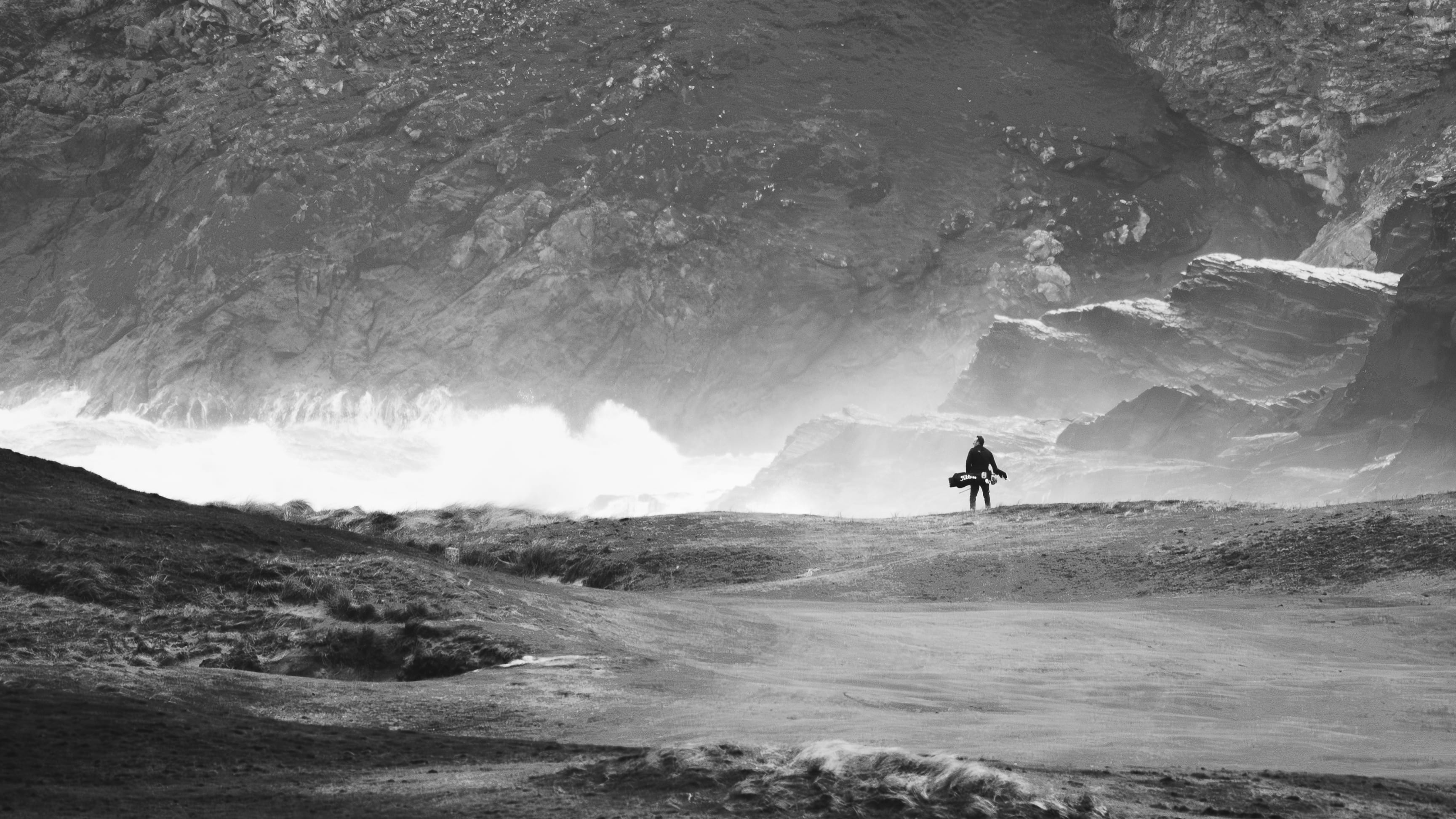 A golfer silhouetted against crashing waves at the base of the cliffs, in black and white