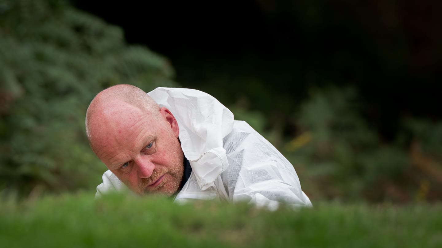 A greenkeeper in white overalls crouched low behind a hedge